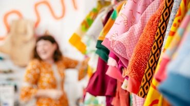 Colourful clothes hanging in a store, with a brunette woman wearing an orange dress, blurred in the background.
