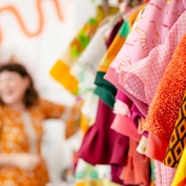 Pink, orange, blue and green colourful clothing hanging on racks in a shop, with a brown haired lady blurred in the background wearing an orange dress